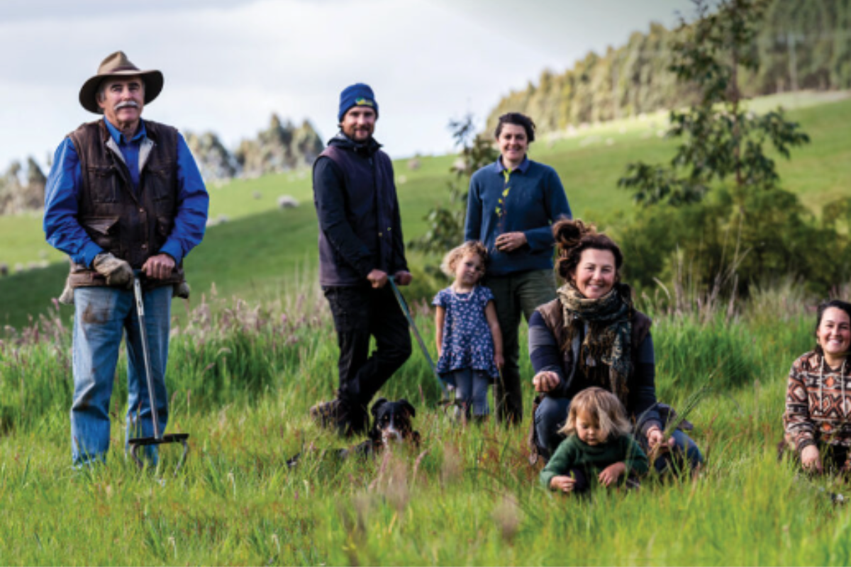 A group of people, including adults and children, standing and kneeling in a grassy field, with a dog at their feet, all participating in land restoration work.